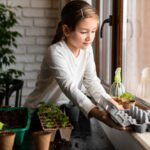 Menina sorridente planta sementes em caixas de ovo, e vasos ecológicos, próxima a uma janela. Com suas mãos na terra, está dando início a um jardim dentro de casa.
