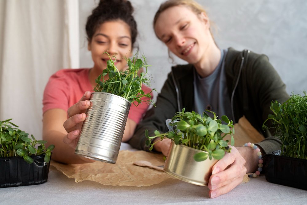 Duas mulheres segurando latas de metal recicladas com plantas dentro, construindo uma horta funcional em apartamento, sorrindo e posando para a foto em um ambiente interno e com luz natural.