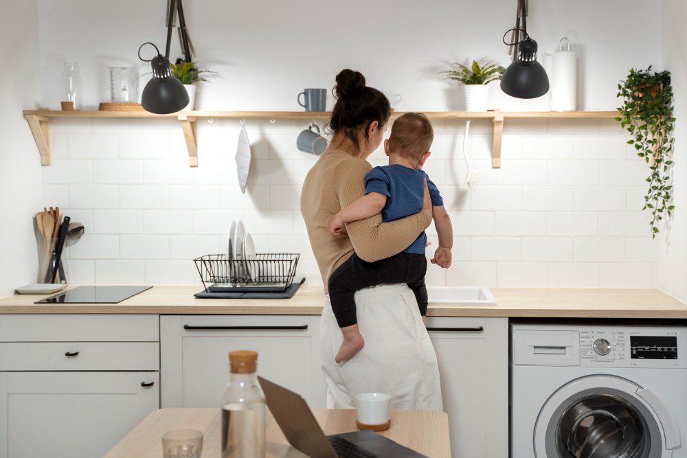 Mulher segurando um bebê em uma cozinha, com utensílios e móveis visíveis ao fundo, transmitindo um ambiente acolhedor.